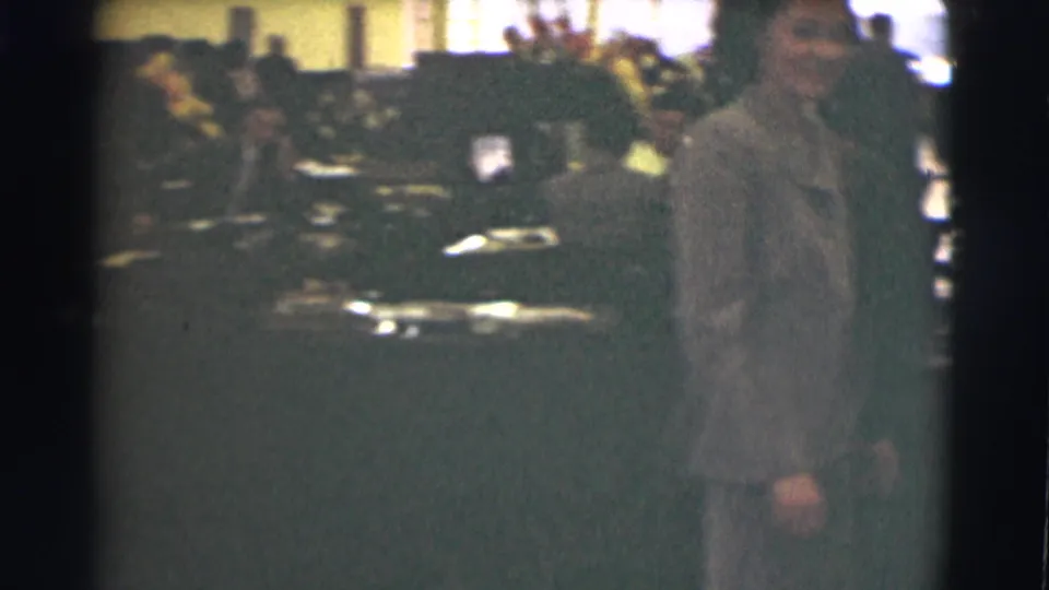 A close view shows the woman in a gray suit near the camera, with office tables, papers, and people behind her.