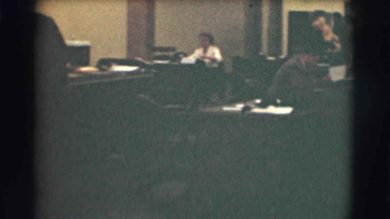 Office interior with desks, papers, a seated worker, and a man standing near the foreground.