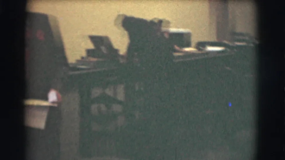 Closer 8mm view of a worker at a desk in the 1959 office, with papers and dark office equipment visible.