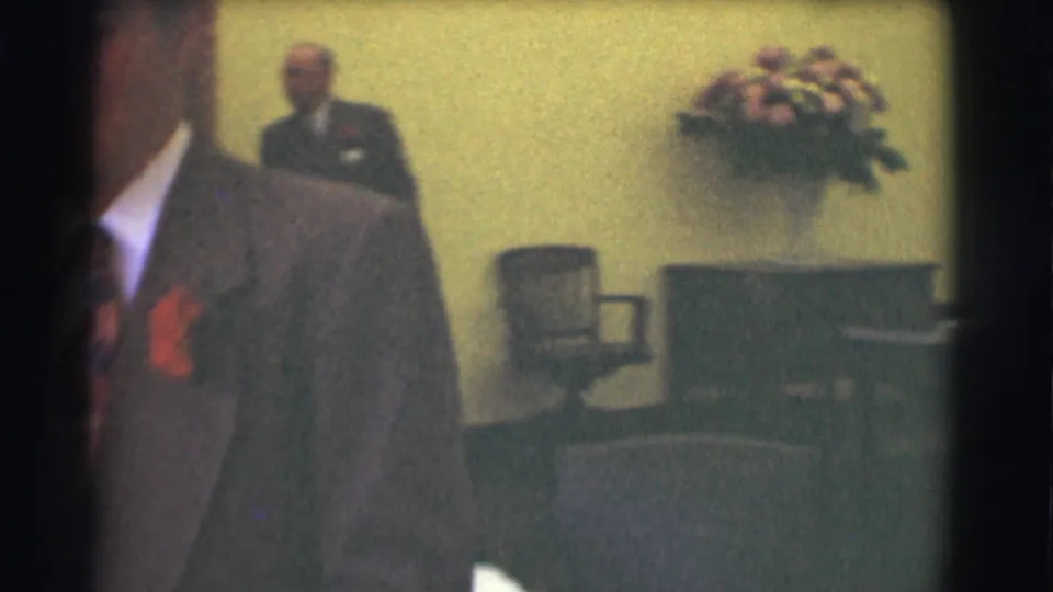 Two suited men appear in a Saint Louis office, with chairs, a desk, and a wall-mounted floral arrangement behind them.