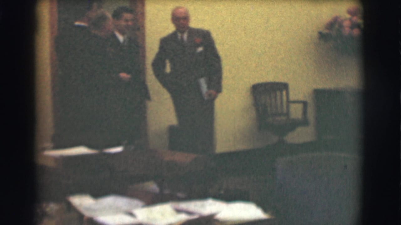 Men in suits stand and sit in a living room with a chair and papers visible.