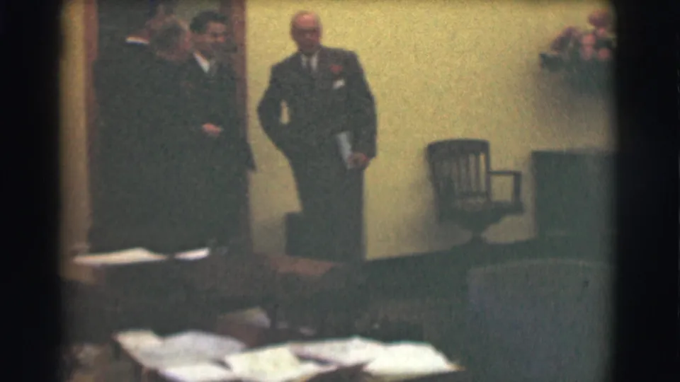 1959 Saint Louis home-movie frame showing suited men in a private living room, with one man standing near a chair and papers in view.