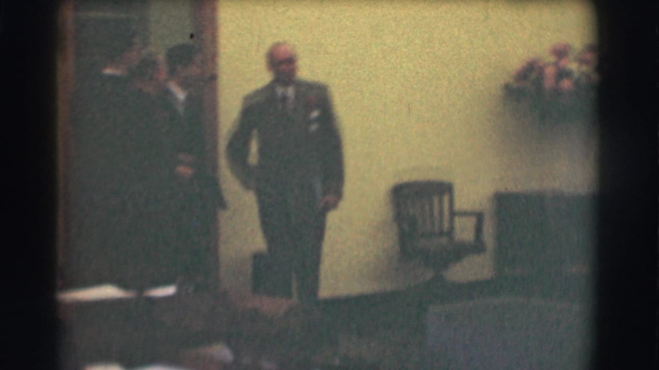 1959 Saint Louis interior frame with a suited man standing near seated or standing adults, a chair, and a softly lit wall.