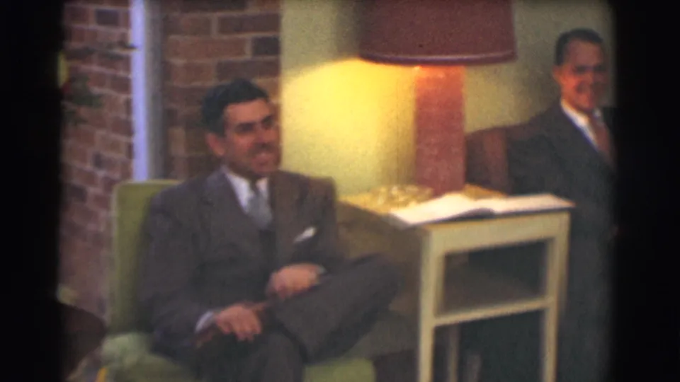 1959 Saint Louis home-movie frame showing suited men seated beside a lamp, side table, green chair, and brick wall accent.