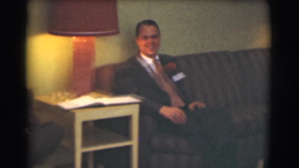 1959 Saint Louis living-room frame showing a suited man seated on a striped couch beside a lit lamp and side table.