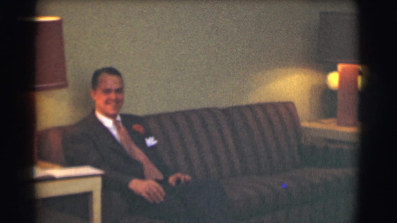 A man in a dark suit and tie sits on a striped couch in a dimly lit interior, consistent with the 1959 Saint Louis funeral-home setting described in the clip metadata.