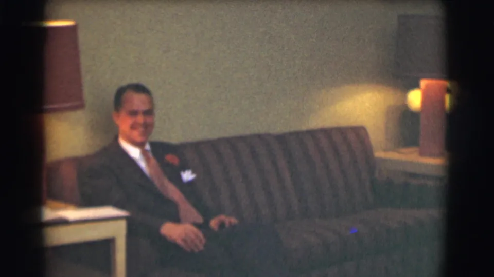 A man in a dark suit and tie sits on a striped couch in a dimly lit interior, consistent with the 1959 Saint Louis funeral-home setting described in the clip metadata.
