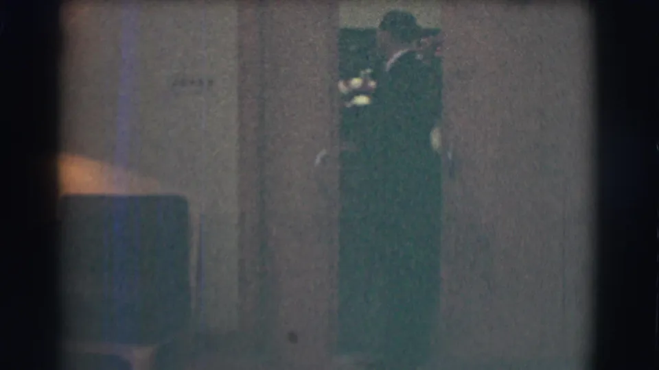 A dark-suited figure stands or passes near a doorway or wall in the dim funeral-home interior, with details partly obscured by grain and exposure.