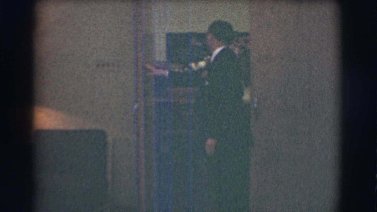Man in a dark suit stands by an open doorway indoors.