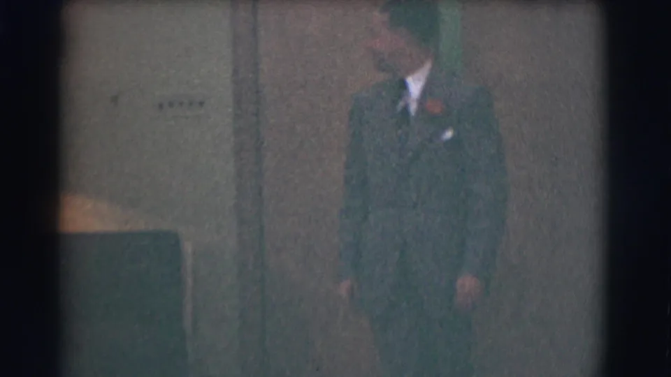 A 1959 Saint Louis frame shows a suited man with a red boutonniere moving through a softly lit interior.