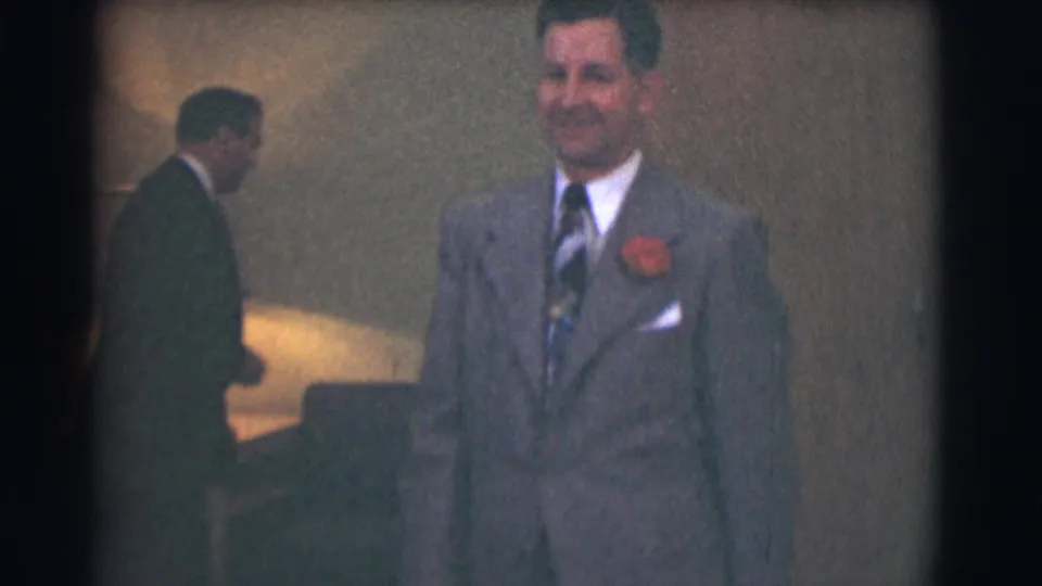 A smiling man in a gray suit and red boutonniere faces the camera in 1959 Saint Louis indoor footage.