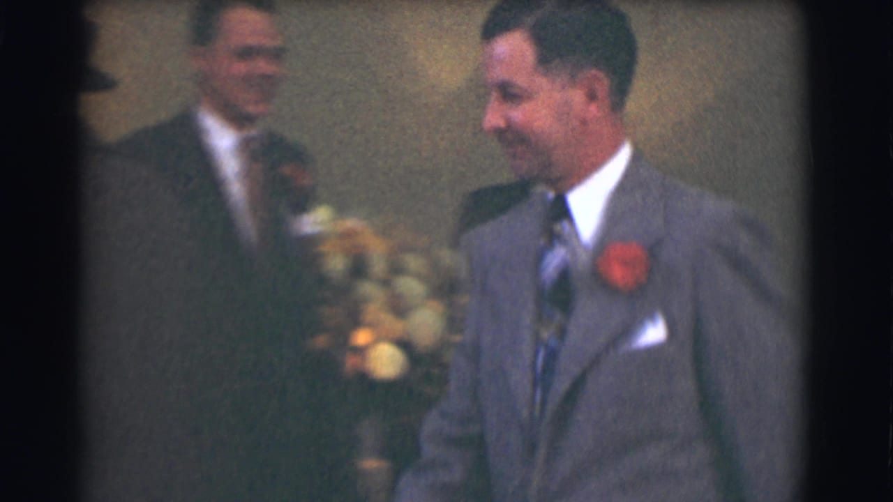 Men in suits stand indoors near flowers, with one man in a gray suit wearing a red corsage.