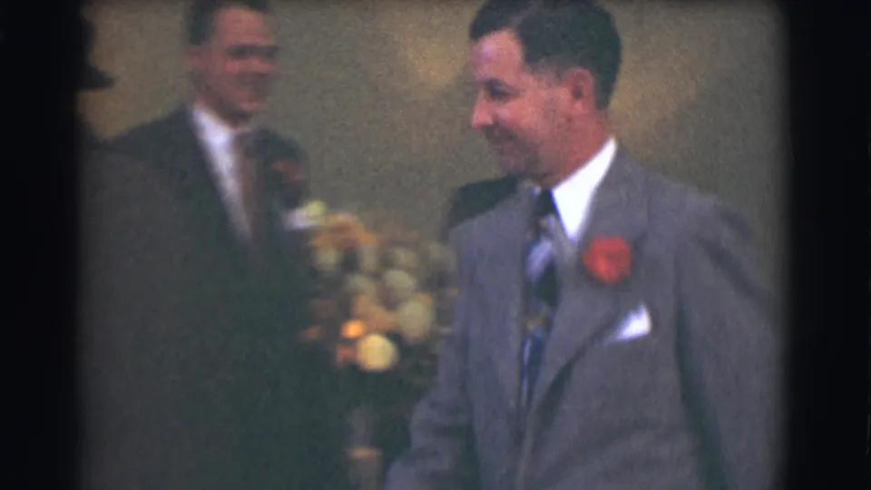 In 1959 Saint Louis home-movie footage, formally dressed men stand indoors as a man in a gray suit with a red corsage smiles.