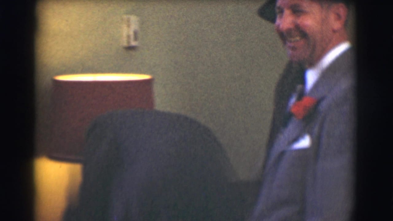 A smiling man in a gray suit stands beside a lamp and upholstered chair indoors.