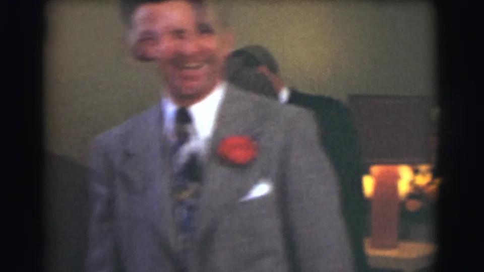 The man in the gray suit laughs toward the handheld 8mm camera, with a red corsage and pocket square visible.