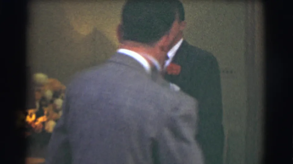 The camera catches the gray-suited man from behind as he turns toward a formally dressed man in a dark suit and red corsage.