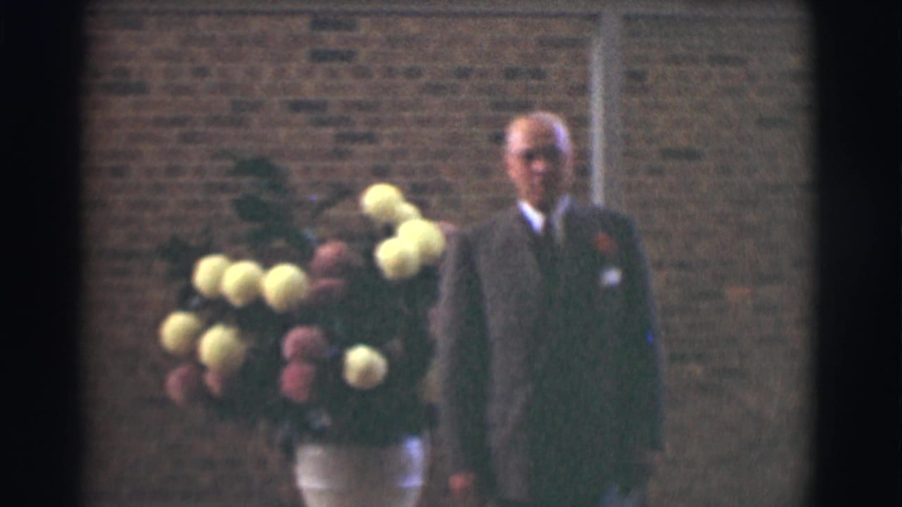 In the Saint Louis church footage, a suited man stands beside a large yellow and pink floral arrangement in a white vase against a brick wall.