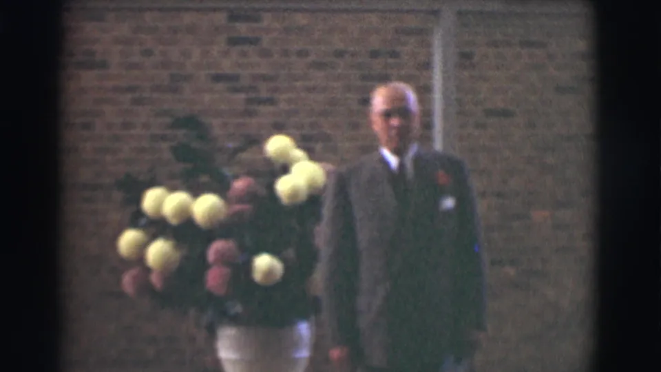In the Saint Louis church footage, a suited man stands beside a large yellow and pink floral arrangement in a white vase against a brick wall.
