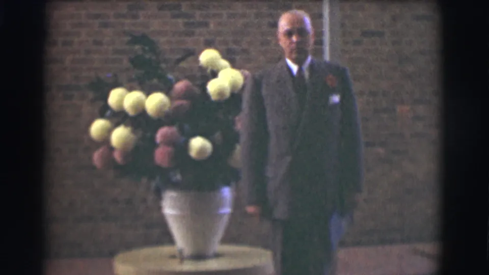 A suited adult appears beside a white vase holding yellow and pink flowers, with the brick church wall visible behind him.