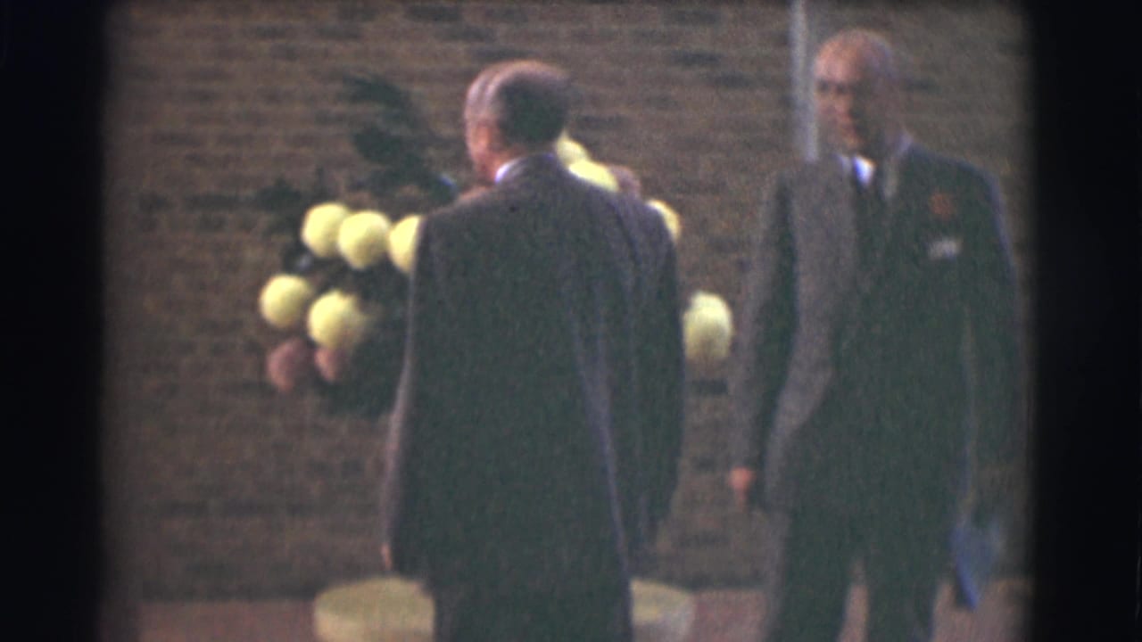 Two suited men stand near a large flower arrangement against a brick wall.
