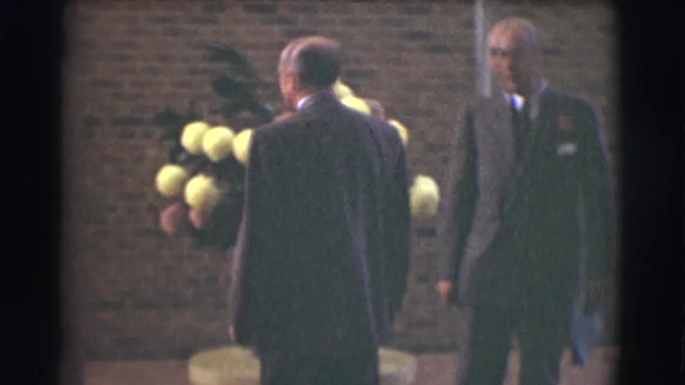 In 1959 Saint Louis home-movie footage, two formally dressed men stand beside a large floral arrangement in front of a brick wall.