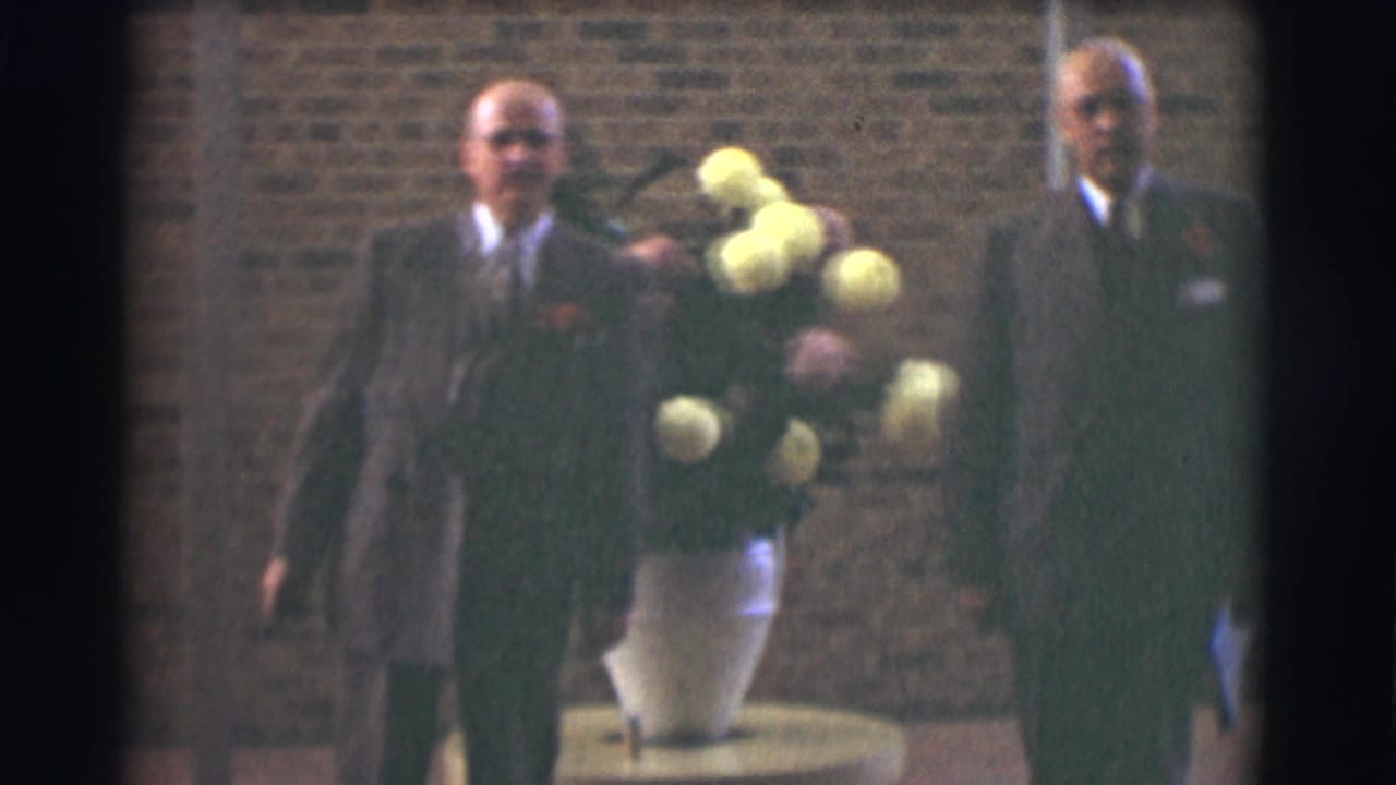 Two men in suits face the camera beside a tall vase of flowers.