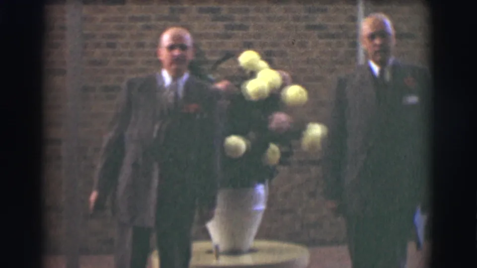 Two older men in suits, one wearing glasses, face the camera beside a large yellow and pink floral arrangement in Saint Louis footage from 1959.