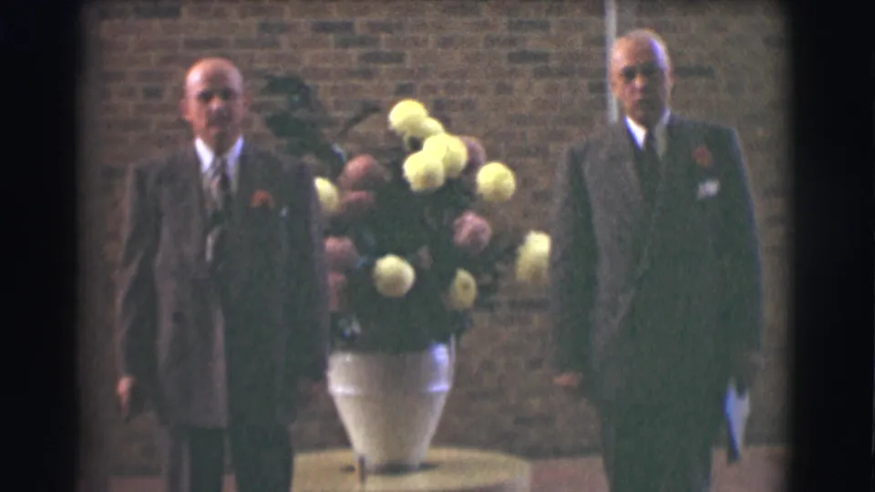 In a soft 8mm frame, two suited men stand on either side of a large white vase with yellow and pink flowers against a brick wall.