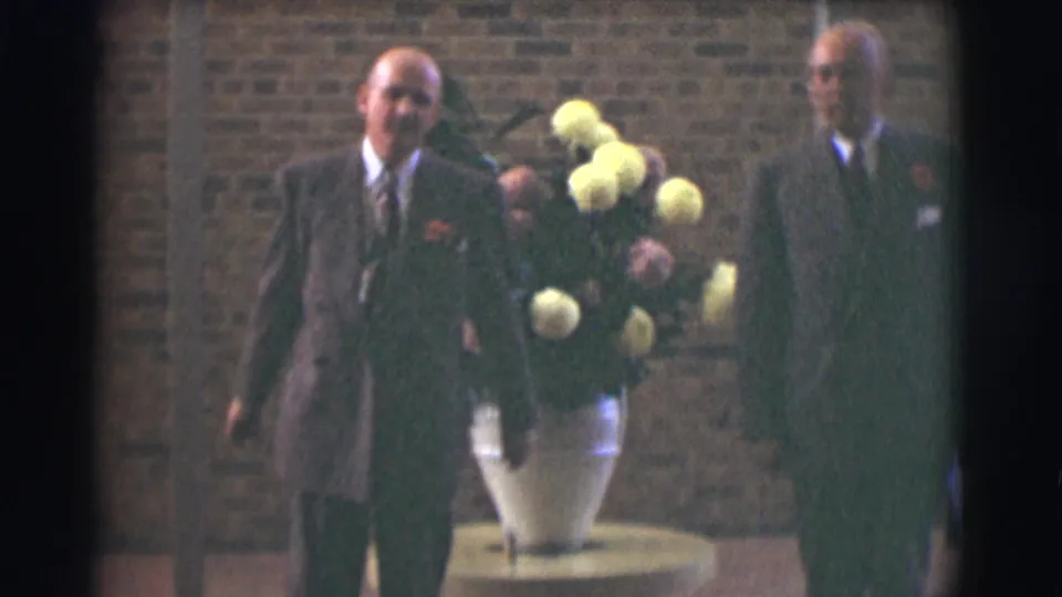 Two formally dressed men hold their pose beside a large floral arrangement in warm, soft 1959 Saint Louis 8mm footage.