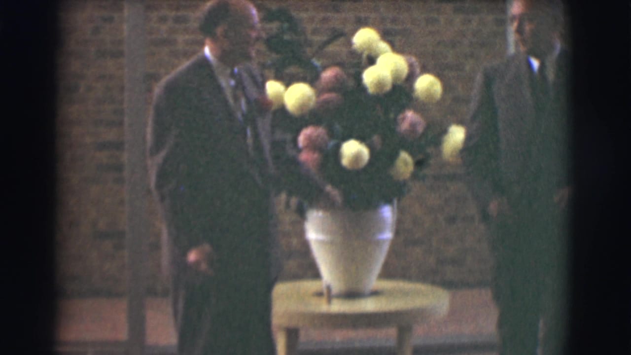 Two men in dark suits stand near a large vase of flowers against a brick wall.