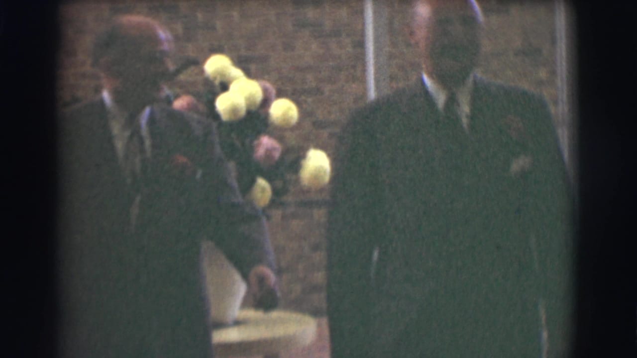 A suited man passes a flower arrangement while another man stands partly in frame.