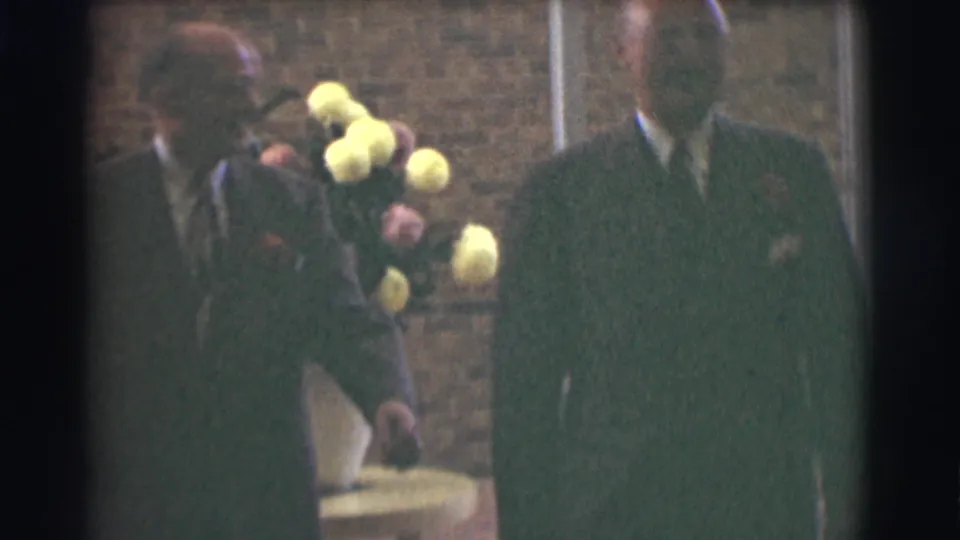 A man in a dark suit walks past the floral display as another suited figure remains partly visible in the 1959 Saint Louis interior.