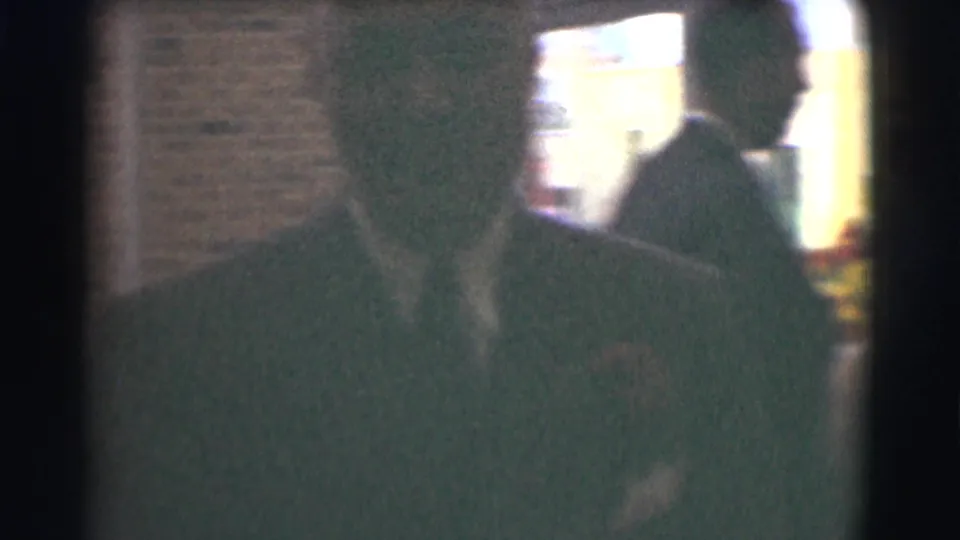 Soft-focus 8mm footage shows a dark-suited man moving close to the camera in the Saint Louis interior, with bright light behind him.