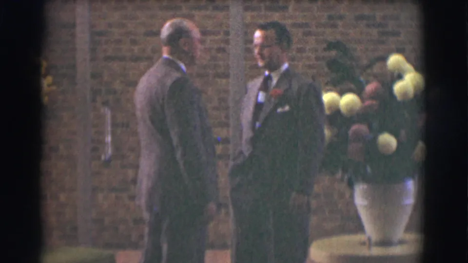 Two adult men in suits face each other near the brick wall and flower arrangement in the 1959 Saint Louis home-movie scene.