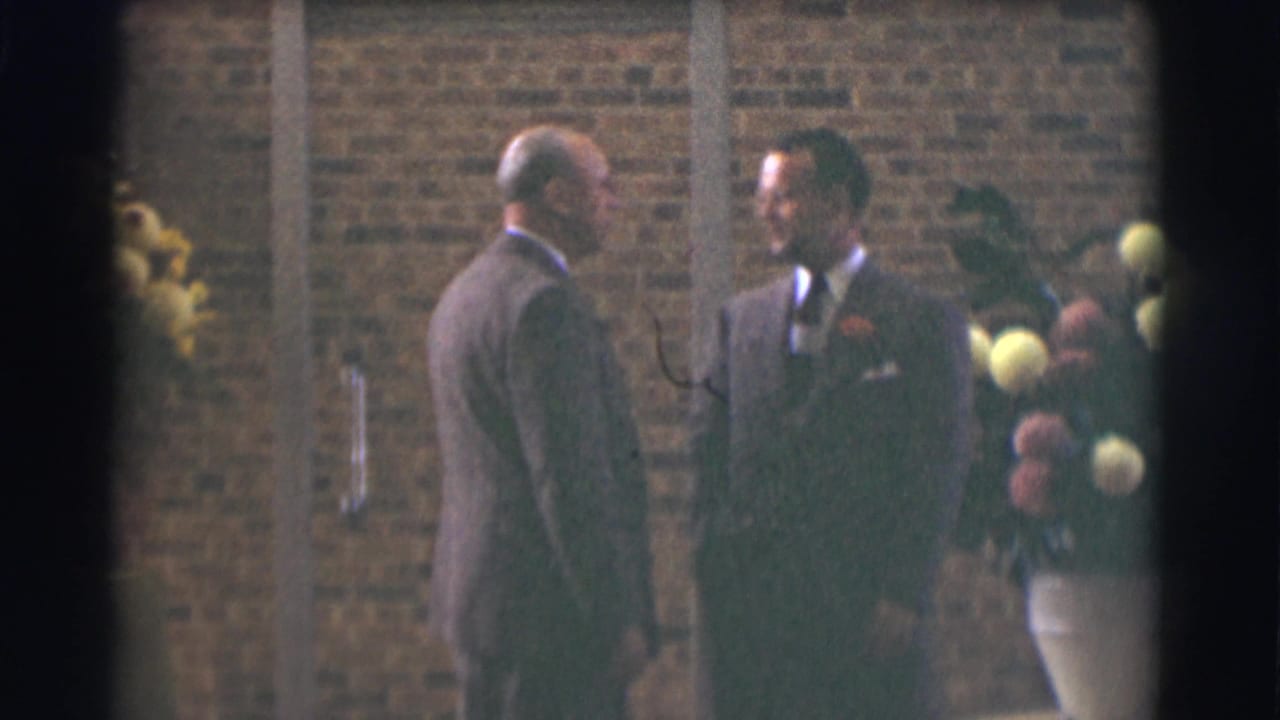 In 1959 Saint Louis 8mm footage, two suited men converse near a brick wall while one holds yellow and pink flowers.