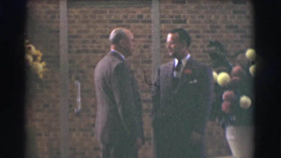 In 1959 Saint Louis 8mm footage, two suited men converse near a brick wall while one holds yellow and pink flowers.