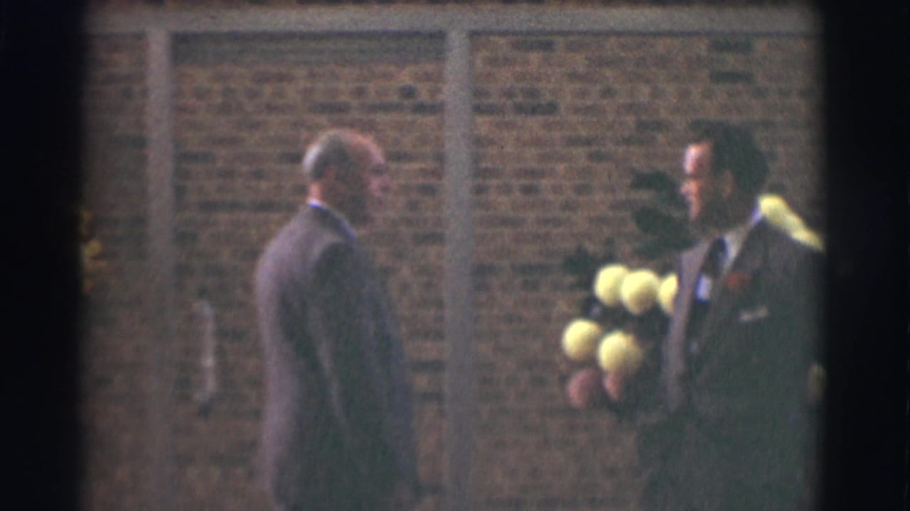 The older man faces the younger suited man with a boutonniere and bouquet outside a Saint Louis brick building in 1959 footage.
