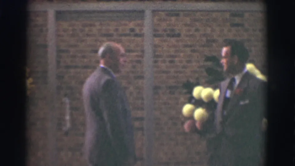 The older man faces the younger suited man with a boutonniere and bouquet outside a Saint Louis brick building in 1959 footage.