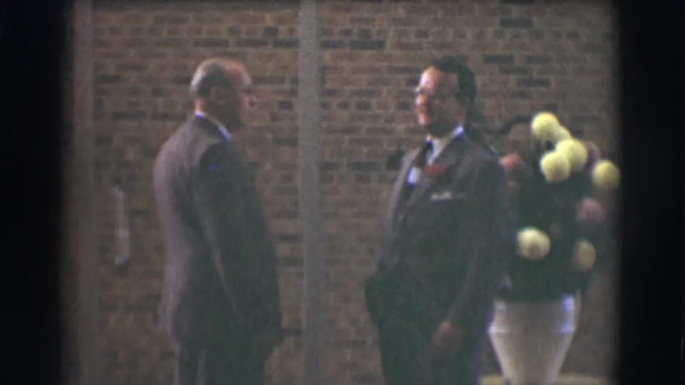 A soft 8mm frame shows two suited men talking outside a brick building in Saint Louis, with flowers at the edge of the scene.