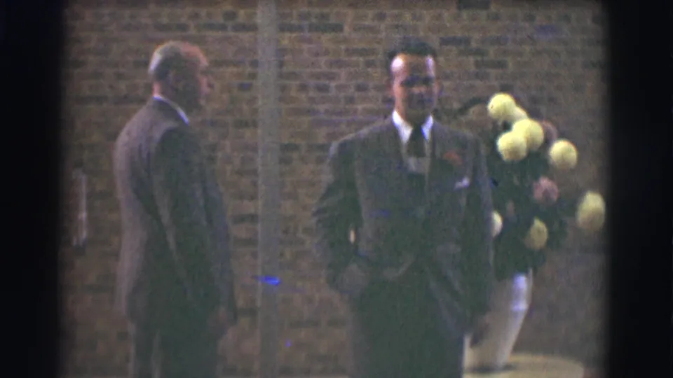 The younger suited man with a boutonniere and bouquet turns toward the camera as the older man remains near the brick wall.