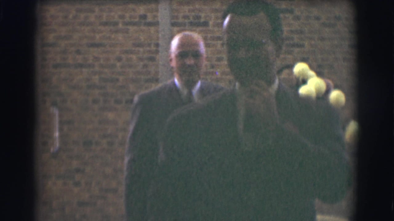 A suited man faces the camera while another dark-coated figure passes in front.