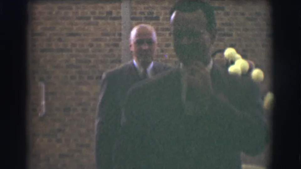 A suited man remains visible near the brick wall as a dark-coated person moves through the foreground.