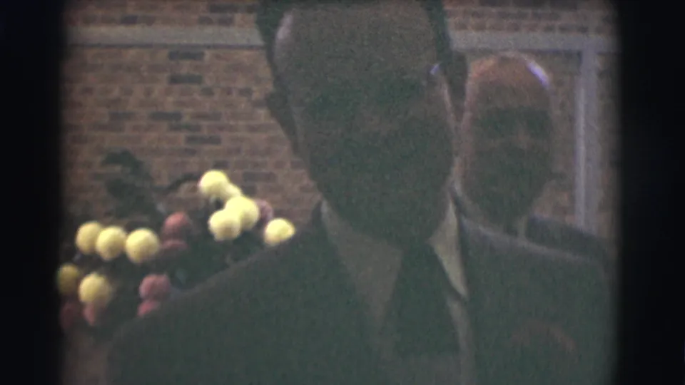 A close, soft 8mm frame shows a man in a dark suit passing the camera with flowers beside him.