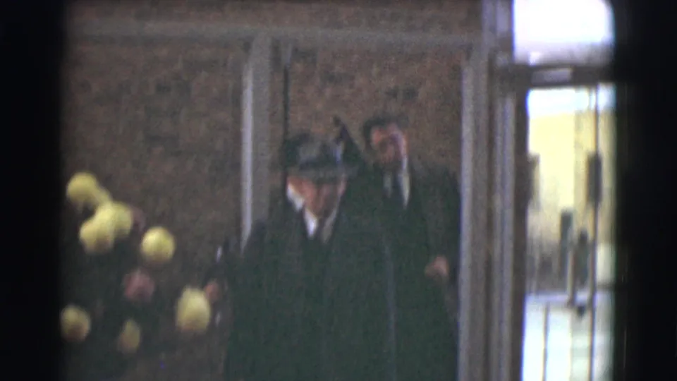 Several dressed figures move near glass doors outside the brick building, with a bouquet beside the entrance.