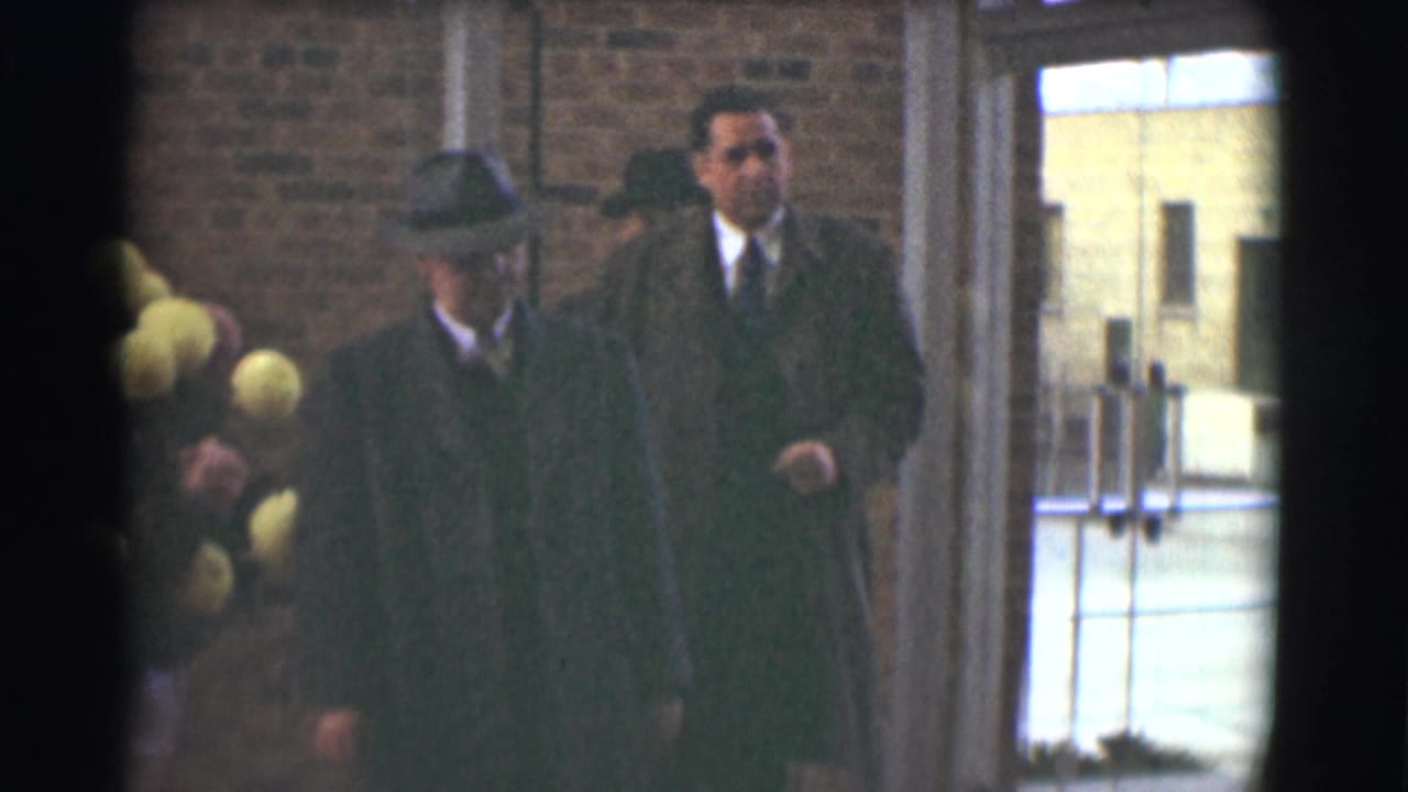 1959 Saint Louis home-movie frame showing suited men in dark coats and hats near a brick entryway during a solemn gathering.