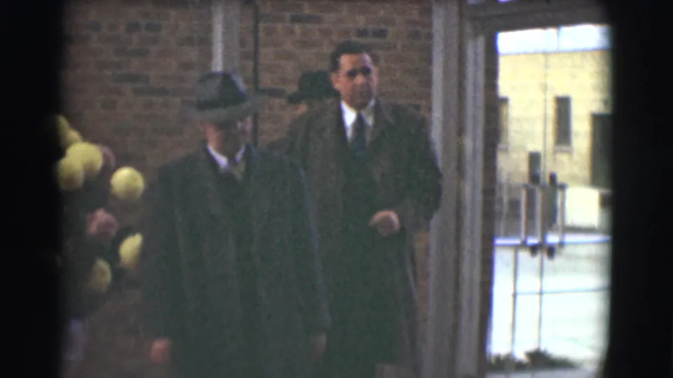 1959 Saint Louis home-movie frame showing suited men in dark coats and hats near a brick entryway during a solemn gathering.