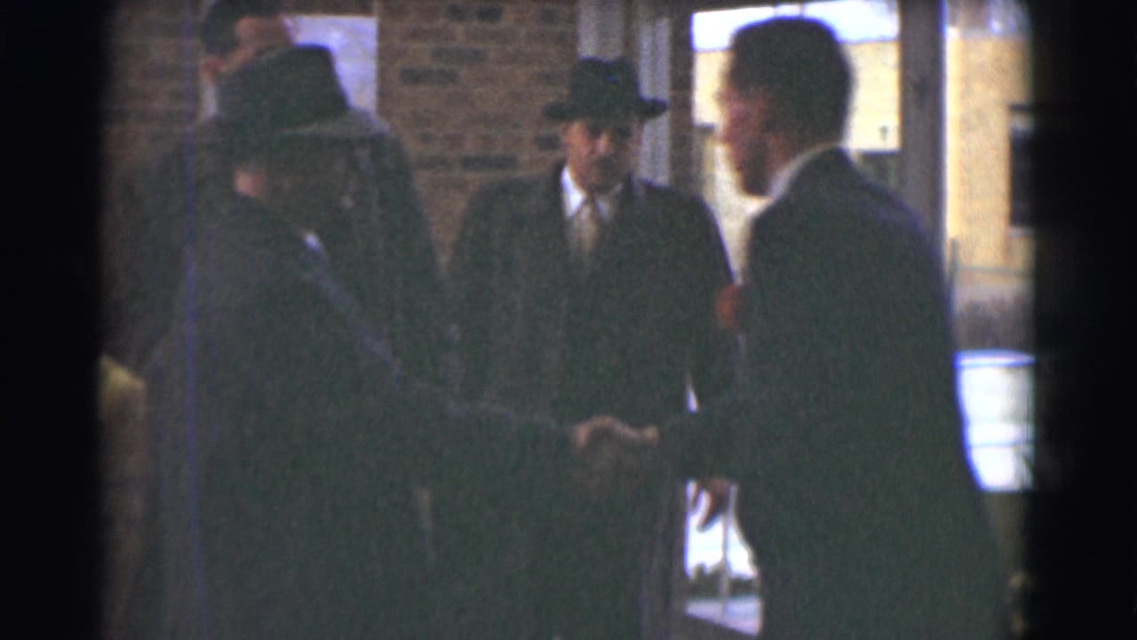 Men in dark formal clothing exchange greetings near a brick doorway in the 1959 Saint Louis funeral scene.