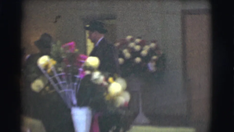Men in dark hats and coats stand near funeral flowers, including a white urn arrangement, in the 1959 Saint Louis footage.