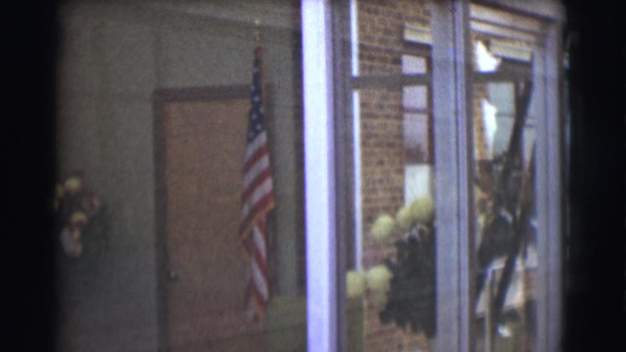 1959 Saint Louis interior frame with a United States flag standing near a doorway, brick wall, window area, and floral arrangements in the background.