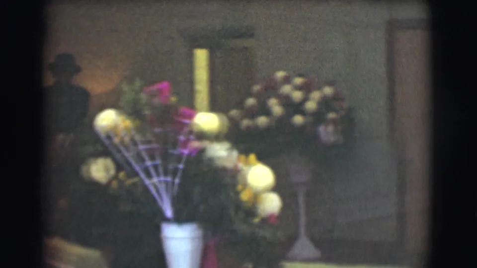 1959 Saint Louis 8mm frame showing a white vase with yellow flowers in the foreground and additional floral arrangements against a dim interior wall.